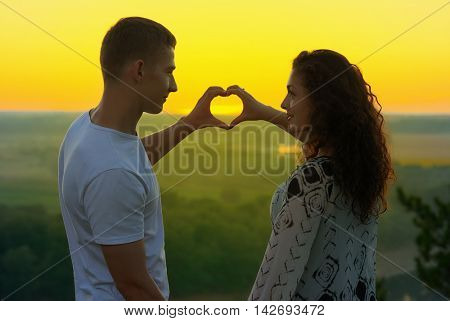 romantic couple at sunset show a heart shape from hands, beautiful landscape and bright yellow sky, love tenderness concept, young adult people