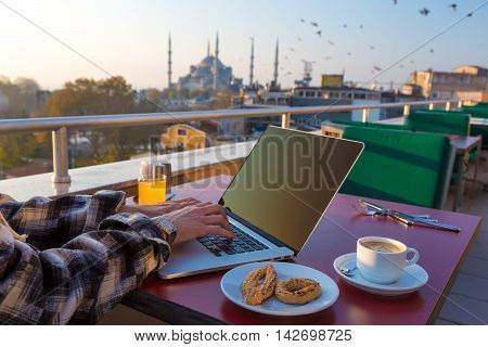 Working Breakfast Person Working on Laptop Computer at Cafe Terrace on Table with Coffee Mug Orange Juice and Bakery Cookies Asian Urban Landscape Mosque