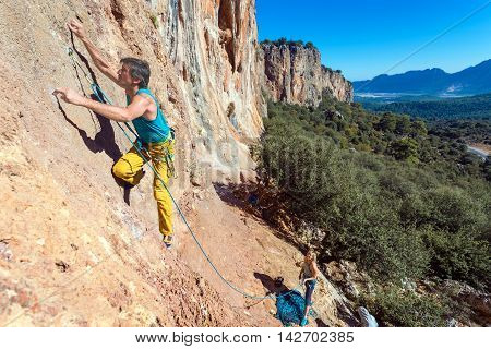 Team of Climbers Man and Woman ascending orange bright rocky Wall with rope and gear Male Leading Female belaying blue Sky and green Forest