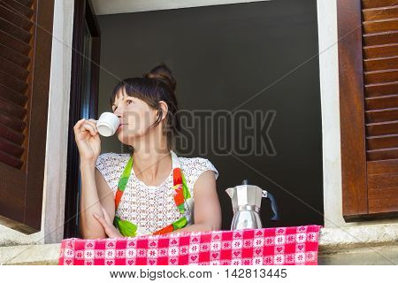Portrait of relaxing young woman with cup of freshly brewed coffee in Italian moka pot and sitting near open window with traditional European wood brown shutters.