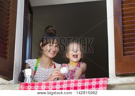 Two years old blonde is having fun with her mother sitting on window sill with white coffee cup and Italian metal moka pot