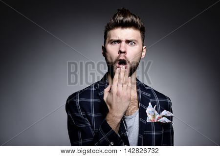 Portrait of young man wriggles in studio over gray background. Hypster gay covers open mouth with his hand. grimanced, fright, fear, surprised, grotesque.