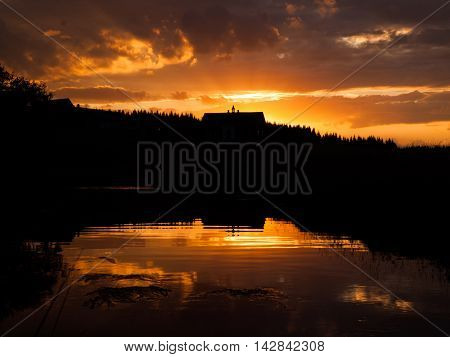 Mountain sunset with river reflection and dark silhouette of forest and rooftop, Jizerka village, Jizera Mountains, Czech Republic