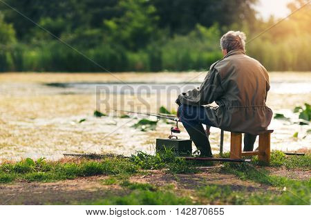 Senior man fishing on a freshwater lake sitting patiently on the shore with his rod and reel as he enjoys his retirement