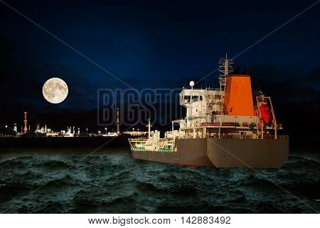 Tanker ship on sea with oil refinery industry in background at night.