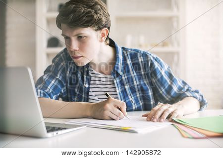 Close up of casual guy working on project at desk with blurry laptop. White brick wall and shelves in the background