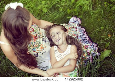Mother and daughter in a beautiful floral sundresses in the summer sit on grass and hug. The girl clung to a pregnant woman and listens to her tummy. The view from the top