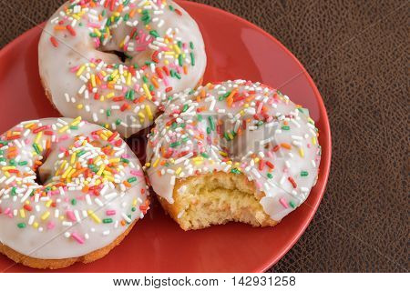 Three white frosted cake donuts with colorful sprinkles, bite taken out of one