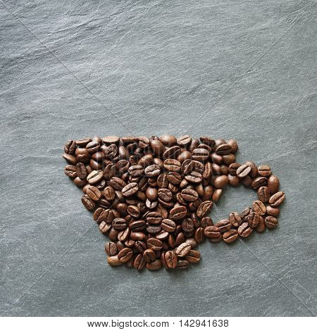 Cup from coffee beans. Heap of roasted coffee bean on grey stone surface texture. Coffee shop or cafe background. Natural stone and cup from beans. Soft color toning, top view
