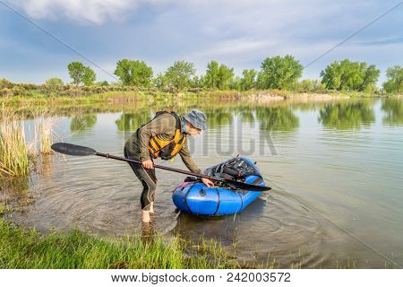 a senior male launching a packraft (one-person light raft used for expedition or adventure racing) on a lake shore in spring scenery