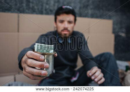Poor And Dirty Man Is Sitting On The Ground And Looking On Camera. He Is Holding A Metal Cup With Do