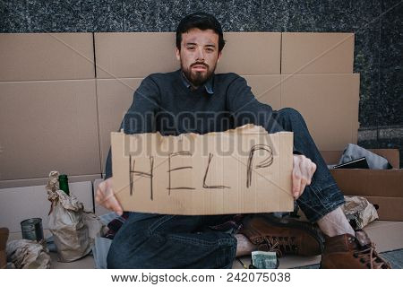 Portrait Of Homeless Guy Sitting On Cardboard And Holding A Help Cardboard In Hands. He Is Looking S