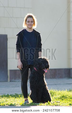 Young Famale Stands With Her Dog On The Green Grass, On The Background Of The Building In The Park, 