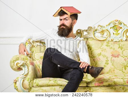 Macho Sits With Open Book On Head, Like Roof. Man With Beard And Mustache Sits On Sofa, White Wall B