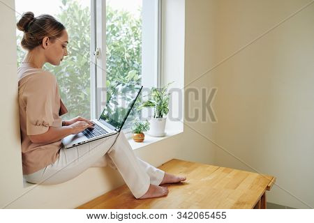 Serious Pretty Young Woman Sitting On Window Sill And Doing Homework For University Online