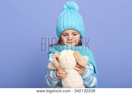Horizontalshot Of Baby Girl Holding Her Soft Dog Toy In Hands And Looking At It, Wearing Warm Clothi