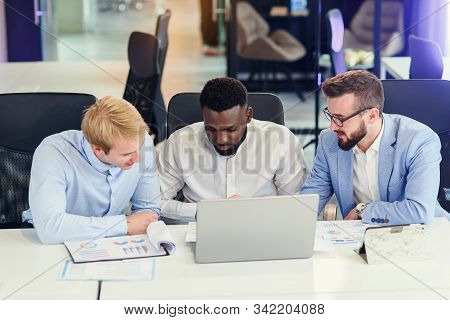 Three Businessmen Sitting At The Modern Office Tables And Analysing Data From Report And Computer To