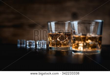 Whisky, Whiskey, Bourbon Or Cognac With Ice Cudes On Black Stone Table And Wood Background