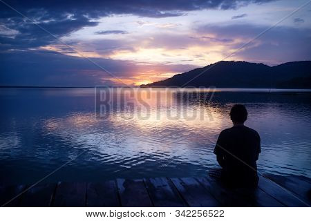 Man Sitting On Dock With Orange Sunset On Dramatic Purple Blue Sky Along Lake Itza, El Remate, Peten
