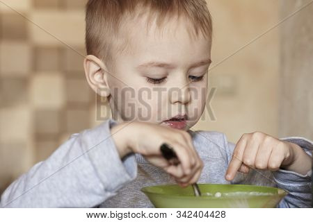 Cute Caucasian Baby Boy With Blond Hairs Points Out With Finger Into The Porridge At The Table, In S