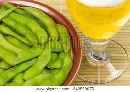 Boiled Edamame In Brown Bowl With Glass Of Beer On Bamboo Mat. Green Soybeans, Maodou, Cooked In Sal