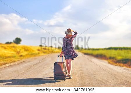 Amazing Woman In Summer Dress And Hat Hitchhiking With Suitcase. Female Walking On The Road With Lug