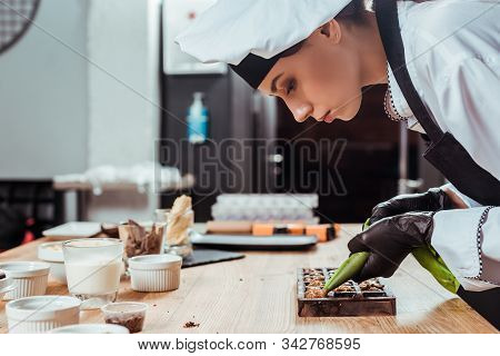 Side View Of Chocolatier In Latex Gloves Holding Pastry Bag With Caramelized Nuts Near Chocolate Mol