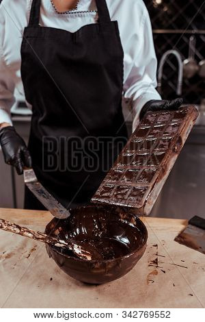 Cropped View Of Chocolatier In Apron Holding Cake Scraper Near Chocolate Molds And Bowl With Melted 