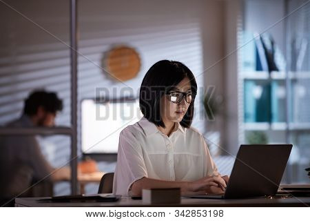 Young Asian Woman In Eyeglasses Sitting At Her Workplace And Typing On Laptop Computer Till Evening 