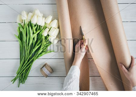 Woman Florist Wrapping Beautiful Bouquet Of White Tulips In Pack Craft Paper On The Wooden Table. Fl