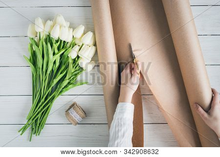 Woman Florist Wrapping Beautiful Bouquet Of White Tulips In Pack Craft Paper On The Wooden Table. Fl