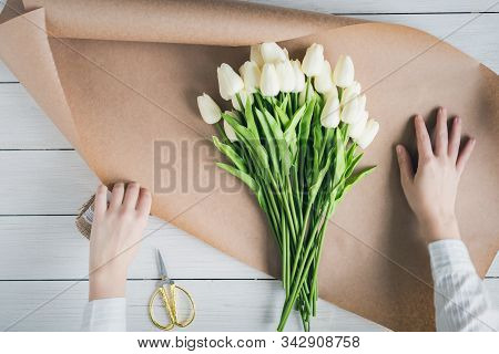 Woman Florist Wrapping Beautiful Bouquet Of White Tulips In Pack Craft Paper On The Wooden Table. Fl