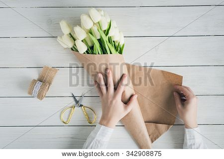 Woman Florist Wrapping Beautiful Bouquet Of White Tulips In Pack Craft Paper On The Wooden Table. Fl