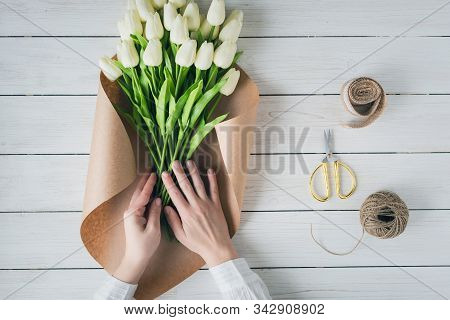 Woman Florist Wrapping Beautiful Bouquet Of White Tulips In Pack Craft Paper On The Wooden Table. Fl