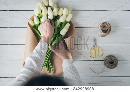 Woman Florist Wrapping Beautiful Bouquet Of White Tulips In Pack Craft Paper On The Wooden Table. Fl