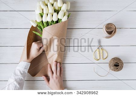 Woman Florist Wrapping Beautiful Bouquet Of White Tulips In Pack Craft Paper On The Wooden Table. Fl