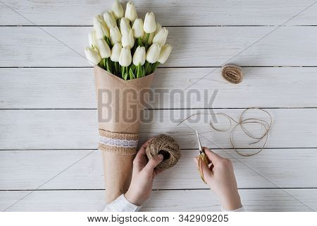 Woman Florist Wrapping Beautiful Bouquet Of White Tulips In Pack Craft Paper On The Wooden Table. Fl