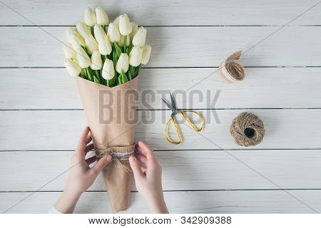 Woman Florist Wrapping Beautiful Bouquet Of White Tulips In Pack Craft Paper On The Wooden Table. Fl