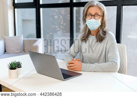 Seious Mature Gray-haired Businesswoman Wearing Mask In Modern Office Looks At The Camera, Typing On