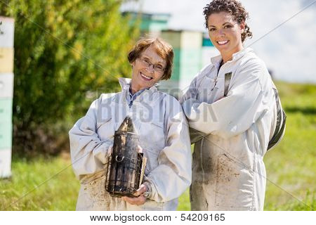 Portrait of confident female beekeepers with smoker standing at apiary