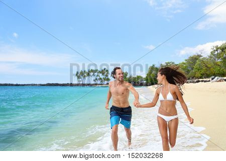 Happy couple laughing together holding hands running having fun splashing water in the ocean waves. Young beautiful fit slim body people enjoying their happy lifestyle in paradise destination beach.