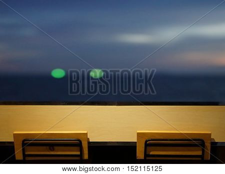 wooden long bar table top with 2 simple chairs at window with seascape at night with bokeh light from fisherman boat faraway as background