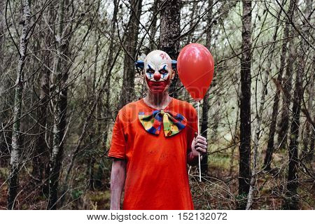 a scary evil clown wearing a dirty costume, holding a red balloon in his hand, in the woods