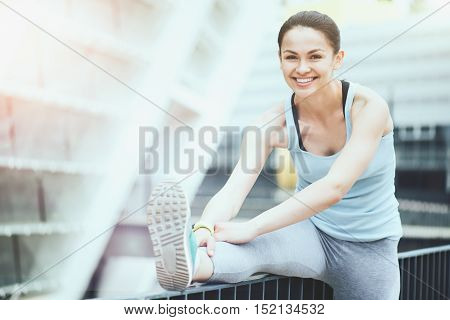 Cheerful exercise. Beautiful slim sporty woman exercising outdoors while stretching and smiling.