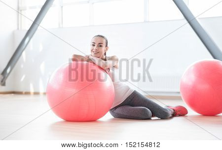 Having a rest. Positive well built young woman lying on the floor and leaning on a fitness ball while being in a gym hall