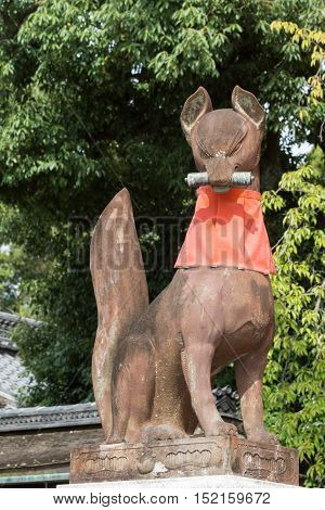 Kyoto Japan - September 17 2016: Fushimi Inari Taisha Shinto Shrine. Kitsune the brown fox messenger and server of God Inari holds a message in its mouth and sports a red apron. Against green tree foliage.
