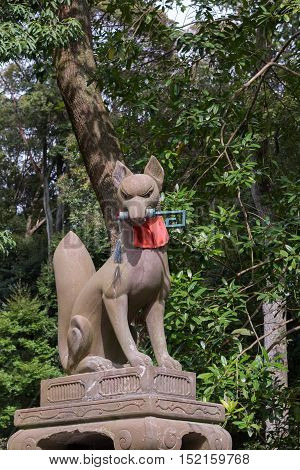 Kyoto Japan - September 17 2016: Fushimi Inari Taisha Shinto Shrine. Kitsune the brown fox messenger and server of God Inari holds the key to good luck in its mouth and sports a red apron. Against green tree foliage.