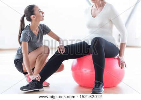 In the right position. Nice attractive skillful coach sitting near the woman and holding her leg while helping her with the exercise