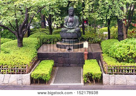 Sitting praying meditating Buddha statute surrounded by lush green foliage of Japanese garden of Asakusa Senso-ji shrine. Tokyo Japan