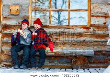 Kids outdoors on beautiful winter day drinking hot chocolate in front of log cabin vacation house